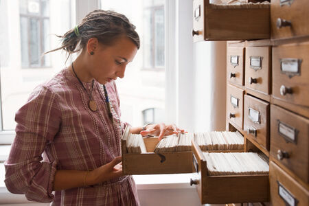 Hipster female student looking through books index archive in library